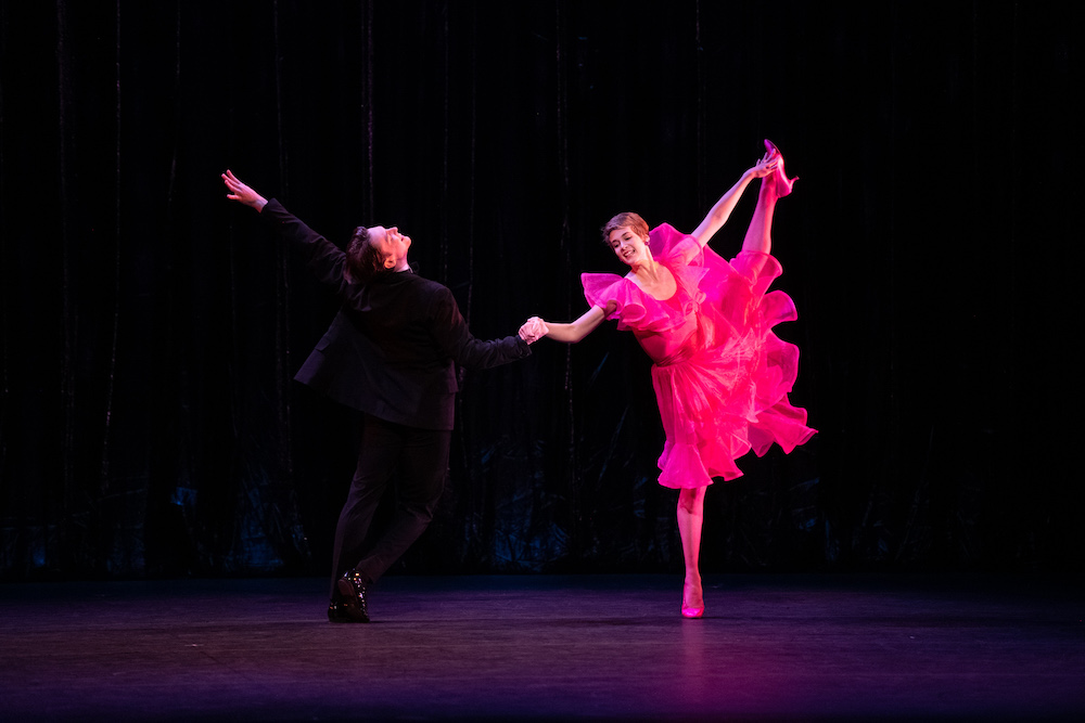 a woman in a flouncy magenta ruffled dress kicks her leg high into the air dancing opposite a gleeful partner in a tuxedo. He is so happy he arches his back and throws out the arm that is not holding hers in an expression of joyous abandon